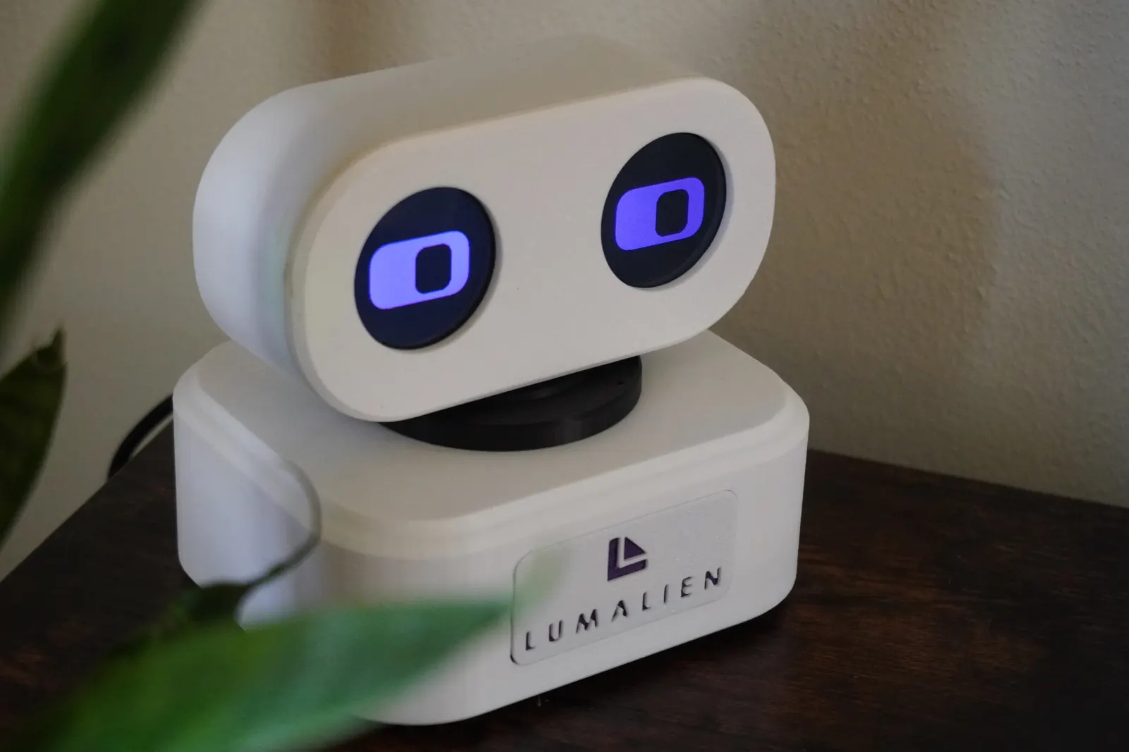 AELX-2 on a dark wooden desk behind a leaf: a white pill-shaped head with violet eyes on a white base with the Lumalien badge